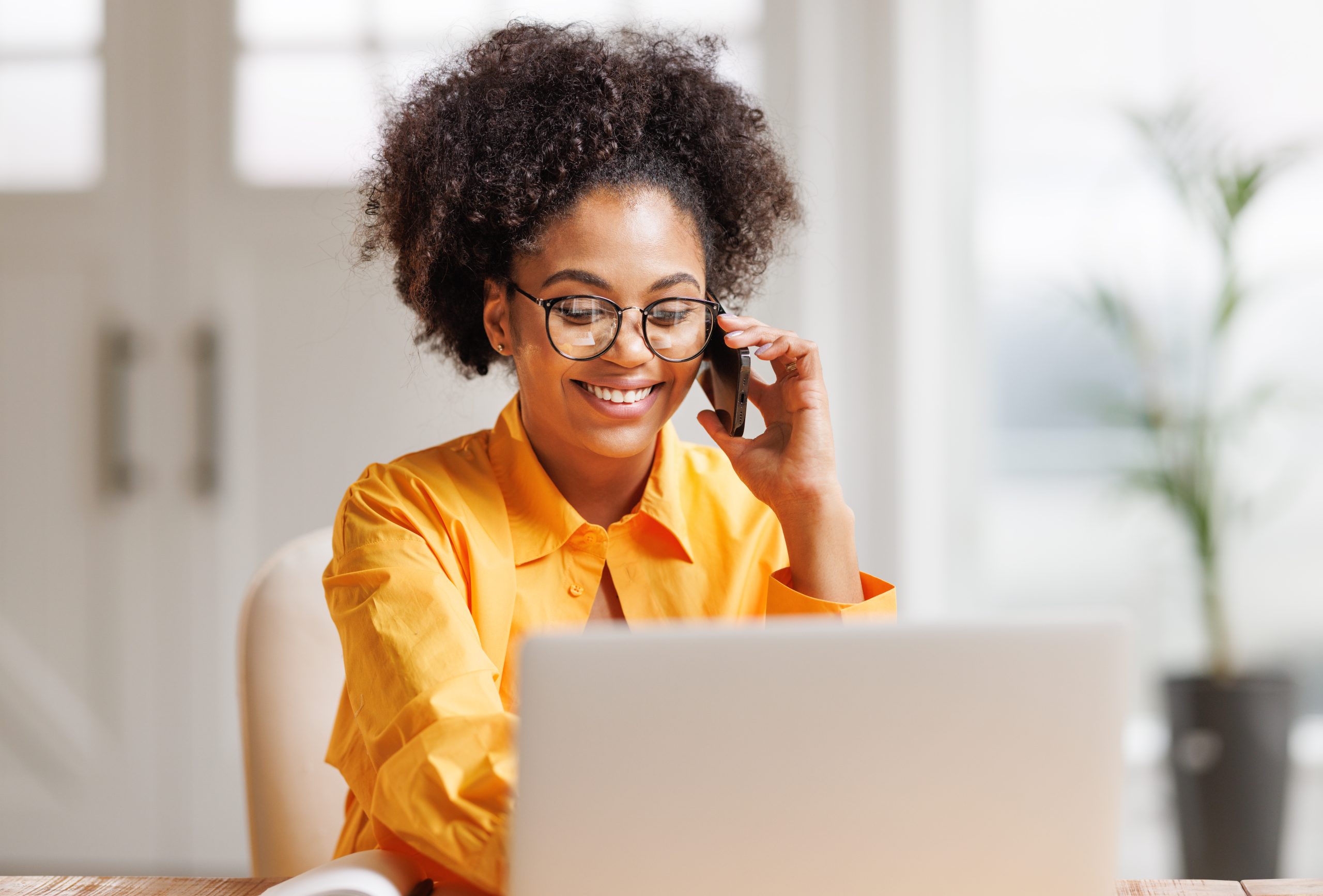 Beautiful young smiling ethnic woman making call via smartphone while working remotely from home while sitting at desk and talking to coworkers